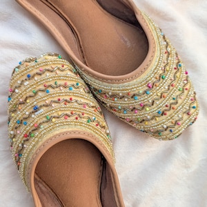 May include: A pair of beige flat shoes featuring intricate gold metallic thread embroidery in a zigzag pattern, accented with small, multicolored beads throughout the upper surface and a smooth, tan-colored interior lining.