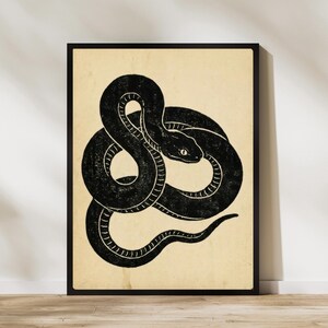 May include: A black and white illustration of a coiled snake in a black frame. The snake has detailed scales and a focused gaze, set against a beige background. The artwork is framed in a simple black border.