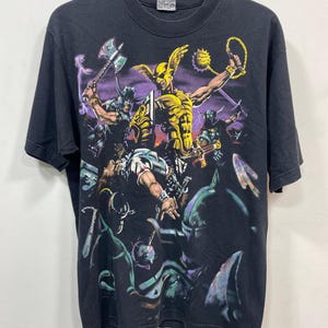Op de afbeelding: Zwart T-shirt met een kleurrijke afbeelding van een gevechtsscène met gouden en paarse figuren. Het ontwerp toont personages met wapens en schilden, waardoor een dynamische compositie ontstaat. Het T-shirt hangt aan een houten hanger.