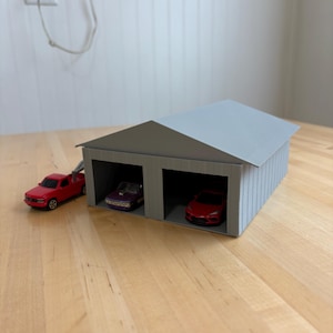 Puede incluir: Un garaje gris en miniatura con dos bahías alberga un coche de juguete morado y uno rojo. Una camioneta roja está estacionada afuera. El garaje tiene un techo corrugado y está sobre una superficie de madera.