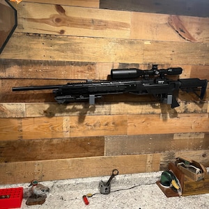 Può includere: Un fucile nero con mirino montato su una parete di legno. Il fucile è montato su una staffa di metallo grigio. La parete è composta da assi di legno di varie tonalità di marrone.