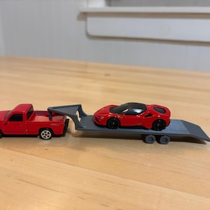 May include: A red toy pickup truck pulling a gray trailer with a red toy sports car. The toy car has a black roof and black wheels. The truck and trailer are on a light brown surface.