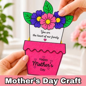 Op de afbeelding: Een handgemaakte Moederdagkaart in de vorm van een bloempot. De roze pot heeft de tekst "Happy Mother's Day" en hartjes. De bovenkant van de kaart heeft paarse en roze bloemen met groene bladeren. De kaart leest "You are the heart of our family."