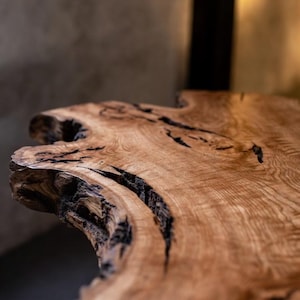 May include: Close-up of a natural wood table surface. The table has a light brown colour with dark brown cracks and knots. The edge of the table has an organic, irregular shape, showcasing the natural grain and texture of the wood.