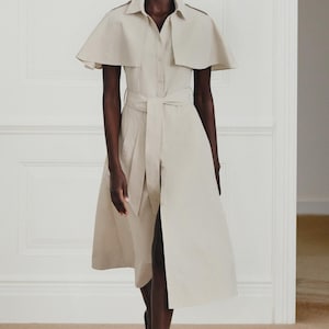 May include: A beige midi-length shirt dress featuring a classic collar, short cape-style sleeves, and a matching fabric tie belt at the waist. The dress has a button-down front closure and a clean, structured silhouette with a flared skirt design.