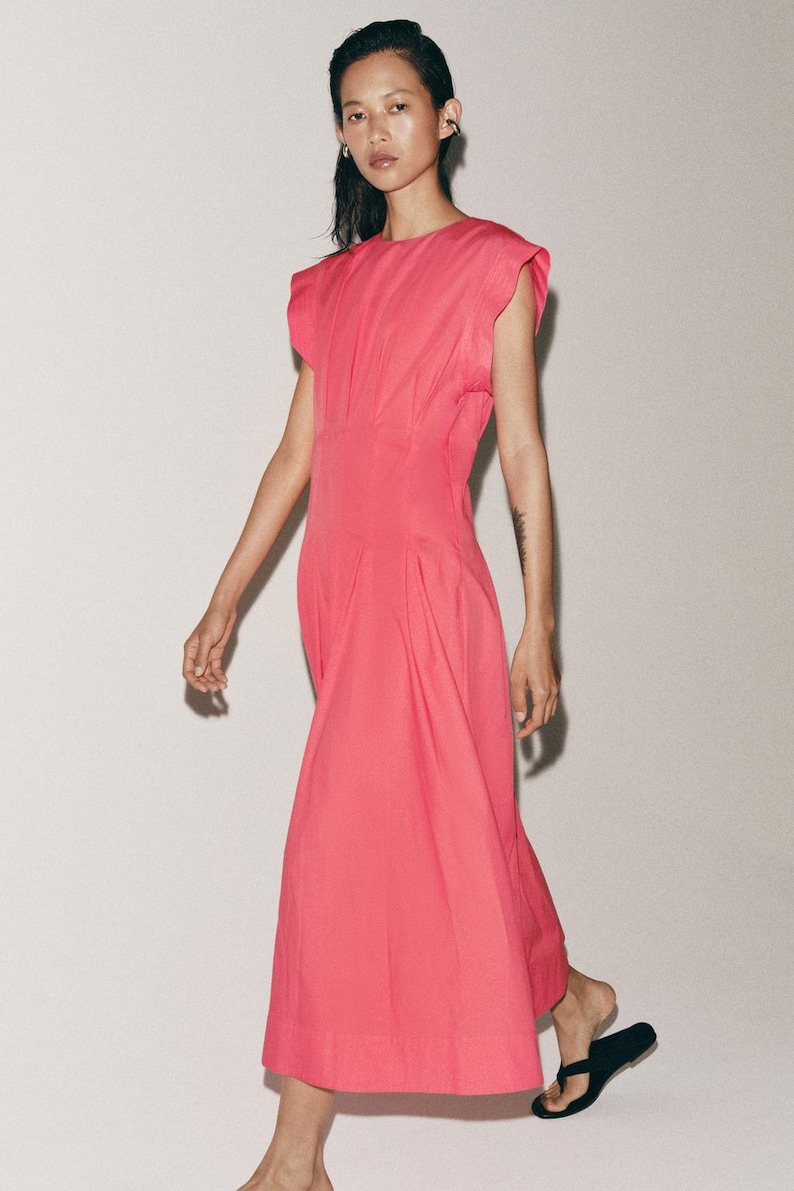 May include: A bright pink midi dress featuring a round neckline, structured cap sleeves, and a fitted waistline that transitions into a pleated skirt. The garment is constructed with a clean, solid-colored fabric and falls to a mid-calf length.