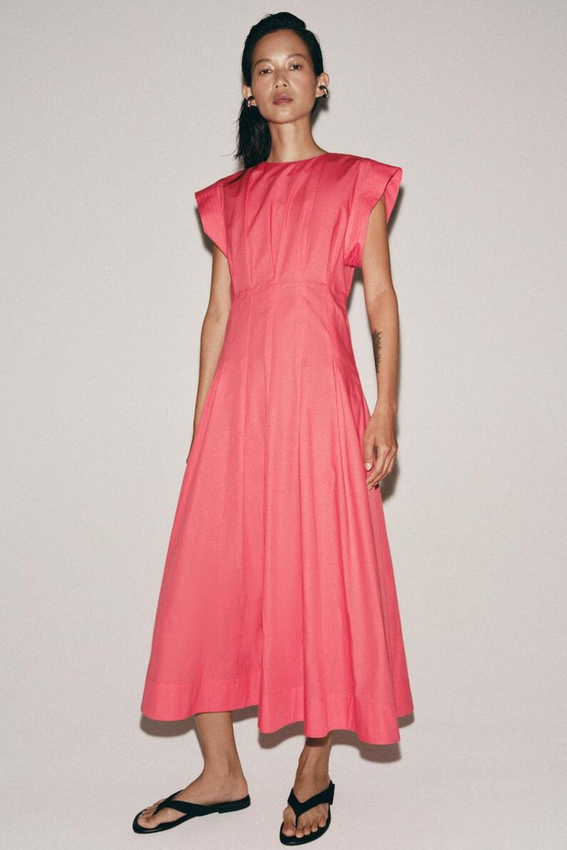 May include: A bright pink midi dress featuring a round neckline, structured cap sleeves, and a fitted bodice that transitions into a pleated, flared skirt. The garment is constructed with vertical seams and falls to a mid-calf length hemline.
