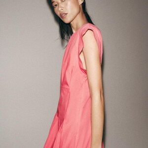 May include: A sleeveless dress in a vibrant shade of pink, featuring a round neckline and structured shoulder detailing. The garment is crafted from a smooth, solid-colored fabric that falls in a clean, straight silhouette along the body.