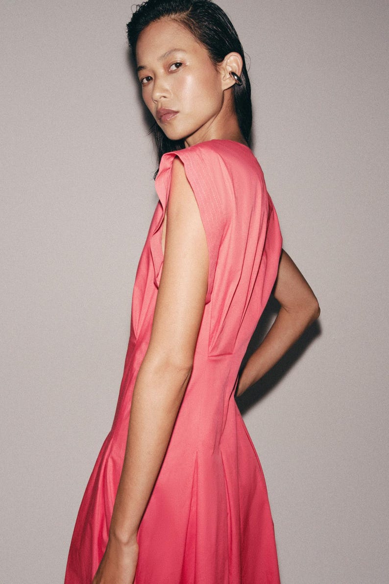 May include: A vibrant pink sleeveless dress featuring structured shoulder details with subtle pleating. The garment has a fitted bodice that transitions into a flared skirt, showcasing a clean, solid color design with a smooth, matte fabric finish.