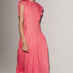 May include: A bright pink midi dress featuring a round neckline, short cap sleeves with subtle ruffles, and a fitted waistline that transitions into a voluminous, pleated skirt that falls below the knees with a clean, straight hemline.