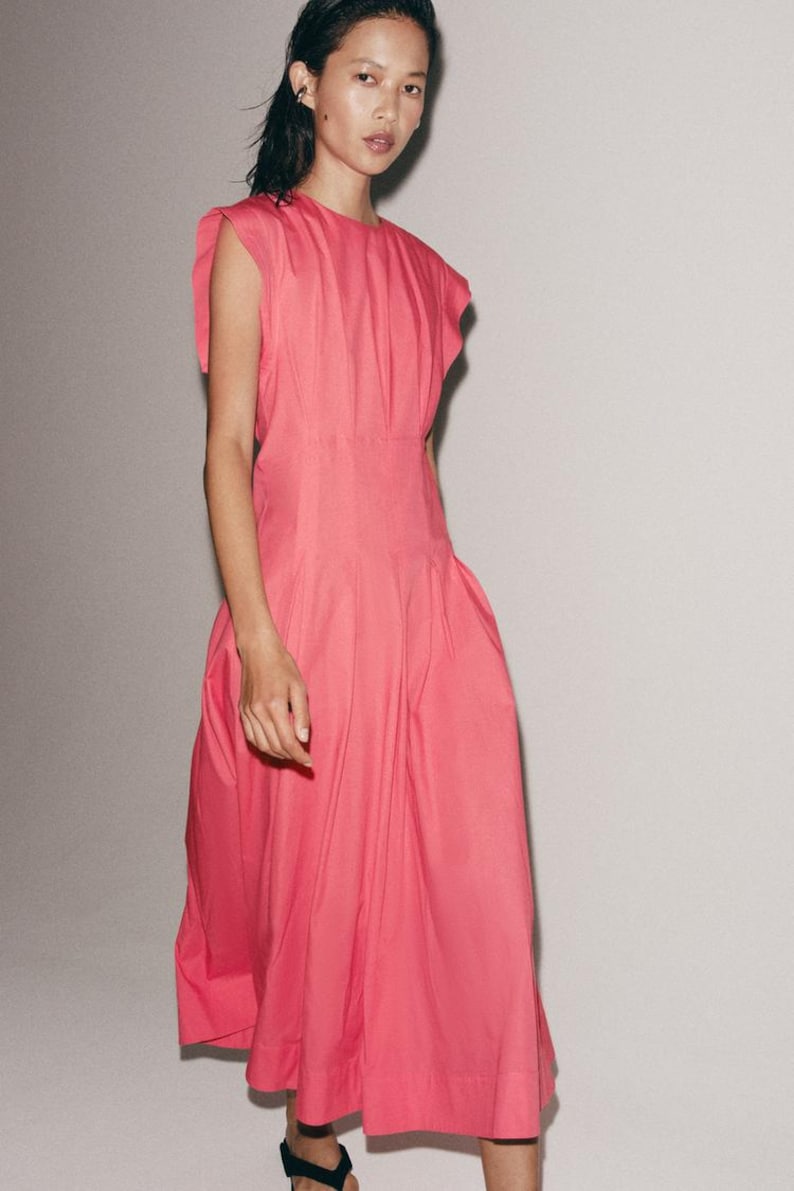 May include: A bright pink midi dress featuring a round neckline, short cap sleeves with subtle ruffles, and a fitted waistline that transitions into a voluminous, pleated skirt that falls below the knees with a clean, straight hemline.