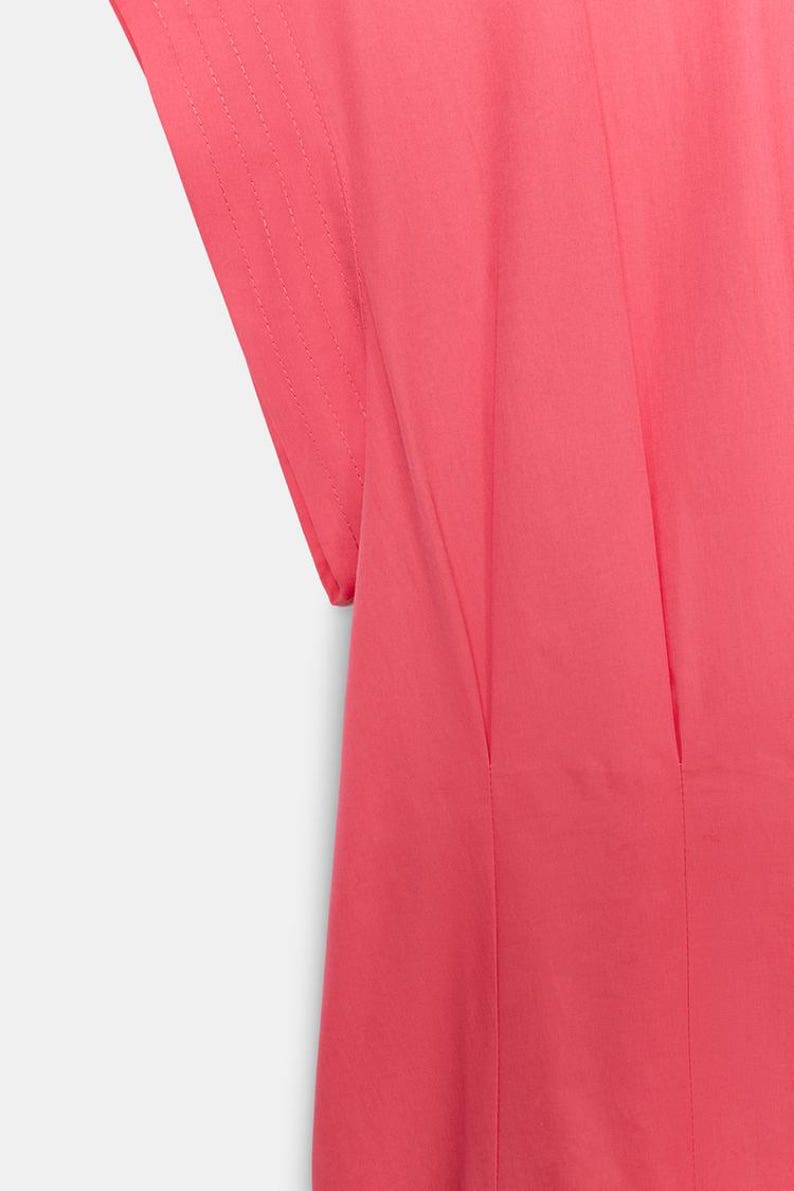 May include: A bright pink garment featuring short sleeves with visible stitched detailing along the edges. The fabric displays vertical pleats or seams that run downward from the shoulder area, creating a structured and clean appearance.