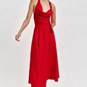May include: A vibrant red midi dress featuring a halter neckline with a draped cowl front, a single shoulder strap secured by a button, and a flared skirt. The dress is paired with simple black thong sandals with red straps.