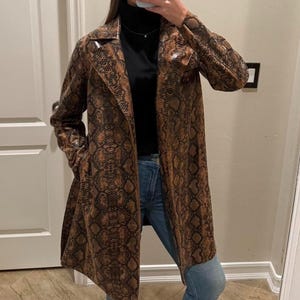 Pode incluir: Um casaco comprido castanho e preto com padrão de pele de cobra. O casaco tem gola de lapela e mangas compridas. O casaco é usado sobre uma gola alta preta e calças de ganga azuis. O casaco é comprido até ao joelho.