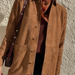 May include: A tan suede jacket with a button-up front, two front pockets, and a collared neckline. The jacket is worn over a burgundy sweater and a patterned scarf. A brown leather shoulder bag is also visible.