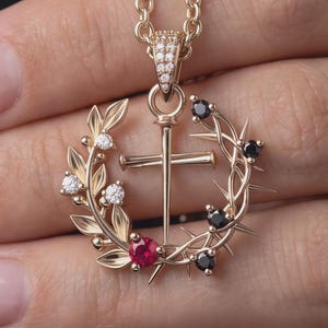 May include: A gold pendant necklace featuring a central cross surrounded by a circular wreath. The left side of the wreath has gold leaves and clear stones, while the right side features thorns and black stones. A single red stone sits at the base.
