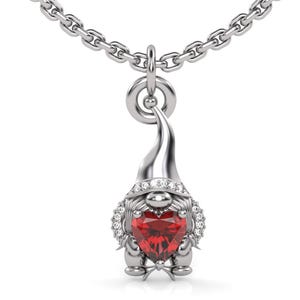 Puede incluir: Un collar con colgante de gnomo plateado. El gnomo tiene una piedra preciosa roja en forma de corazón en el centro y está rodeado de pequeñas piedras preciosas transparentes. El colgante cuelga de una cadena plateada.