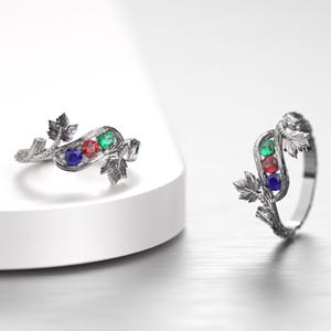 Könnte beinhalten: Zwei silberne Ringe mit einem Blattdesign. Ein Ring liegt, der andere steht. Jeder Ring hat drei farbige Edelsteine: blau, rot und grün. Die Ringe liegen auf einer weißen Oberfläche.