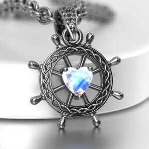 May include: A dark metallic pendant shaped like a ship's wheel with a braided inner ring and a central heart-shaped iridescent moonstone. The pendant hangs from a matching dark metal chain, featuring a small bail adorned with tiny clear stones.