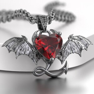 May include: A dark metallic pendant necklace featuring a faceted heart-shaped red gemstone centerpiece. The pendant is flanked by detailed bat-like wings and includes a curved tail element at the bottom, all suspended from a matching dark chain.