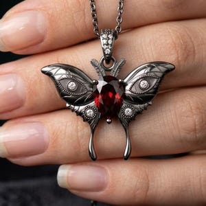 Puede incluir: Un collar con colgante de mariposa plateada con una gran gema roja ovalada en el centro. Las alas de la mariposa están detalladas con patrones grabados y pequeñas gemas transparentes. La cadena del collar también es plateada.