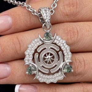 May include: A silver pendant necklace with a circular design, a sunburst centre, and three green gemstone accents. The pendant is encrusted with small, clear gemstones and hangs from a delicate chain.