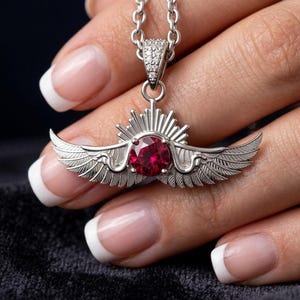 May include: A silver-toned pendant necklace featuring a central round-cut deep red gemstone. The pendant is designed with symmetrical feathered wings extending outward and a sunburst motif above the stone. The bail is encrusted with small clear stones.