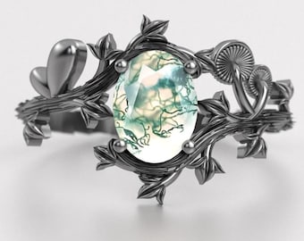 May include: A silver ring featuring an oval-cut gemstone with green and white inclusions. The ring's band is adorned with leaf and mushroom details, creating a nature-inspired design. The ring is set against a white background.