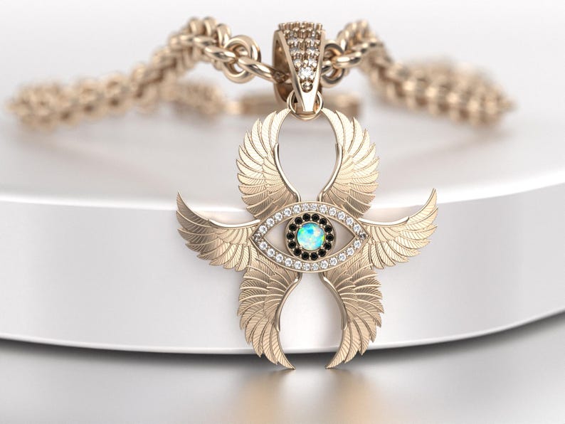 May include: Gold-toned pendant necklace featuring an Ankh symbol with stylized wings and an eye design. The eye has a central blue-green stone surrounded by black and clear stones. The chain is a thick, textured gold. Jewelry.