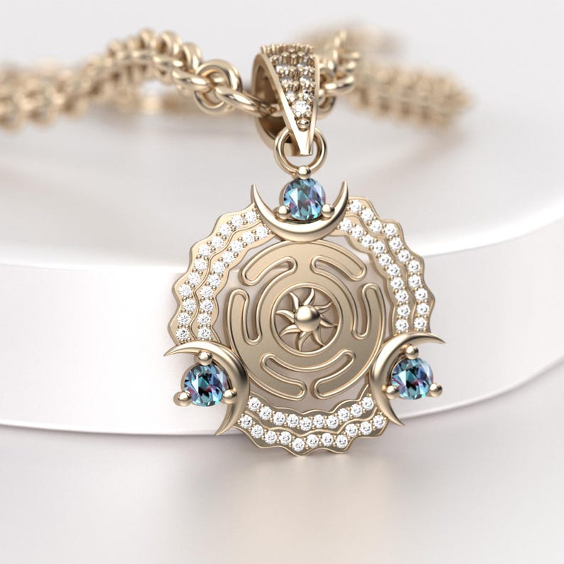 May include: Gold-toned pendant necklace featuring a circular design with a sunburst center and crescent moon accents. The pendant is adorned with small, sparkling gemstones and three blue-green stones. The necklace chain is also gold-toned.