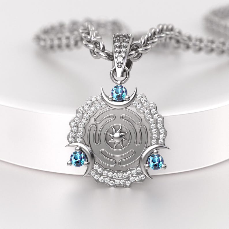May include: Silver pendant necklace featuring a circular design with a sunburst center and a maze pattern. The pendant is adorned with blue gemstones and small clear stones. The necklace has a silver chain.