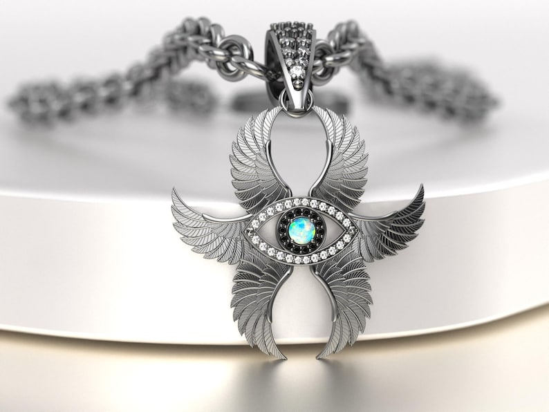 May include: A silver-colored pendant necklace featuring an Ankh symbol with stylized wings. The pendant has an eye design with a central blue-green stone, surrounded by clear and black gemstones. The chain is a thick, textured silver.