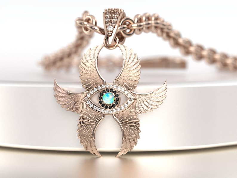 May include: Rose gold pendant necklace featuring an eye-shaped design with feathered wings. The pendant has a central opal-like stone, surrounded by white and black gemstones. The chain is also rose gold.