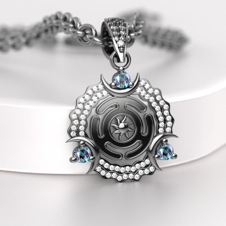May include: A silver pendant necklace featuring a circular design with a sunburst center, adorned with small clear gemstones and three blue-green gemstones. The pendant hangs from a silver chain.