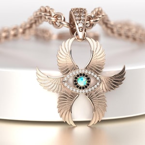 May include: A rose gold pendant necklace featuring a stylized winged eye design. The pendant has an eye-shaped center with a blue-green stone and black accents, surrounded by small clear stones. The wings are detailed with feather patterns.