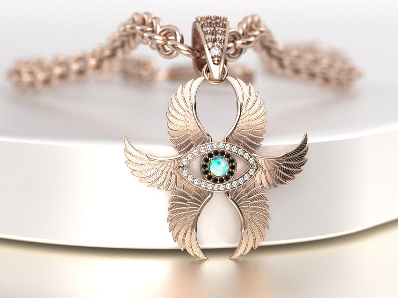 May include: A rose gold pendant necklace featuring a stylized winged eye design. The pendant has an eye-shaped center with a blue-green stone and black accents, surrounded by small clear stones. The wings are detailed with feather patterns.