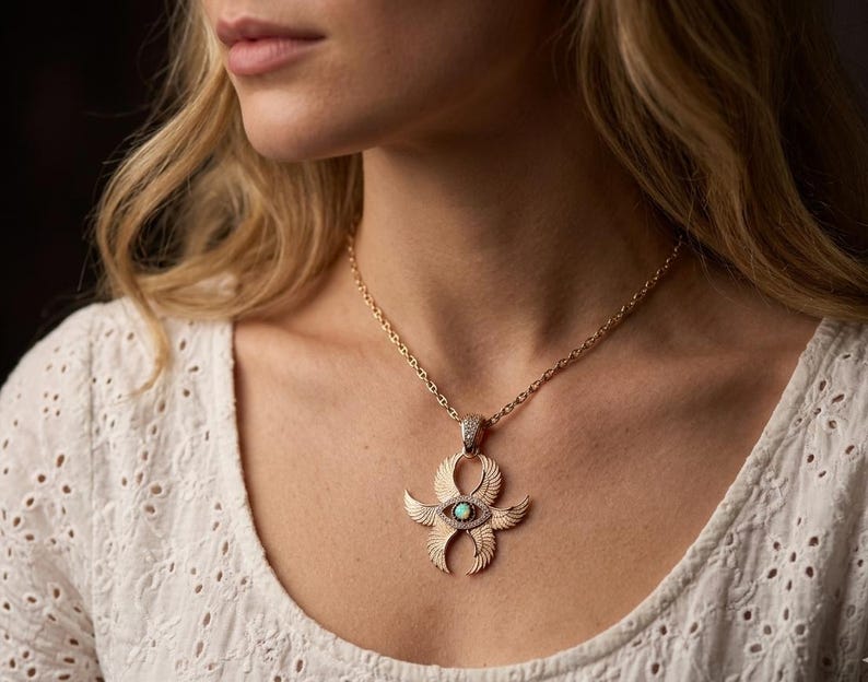 May include: Gold-toned pendant necklace featuring an Ankh design with an eye-shaped opal stone at the center. The pendant has detailed wing-like elements and is suspended from a delicate chain. The necklace is worn against a white eyelet top.