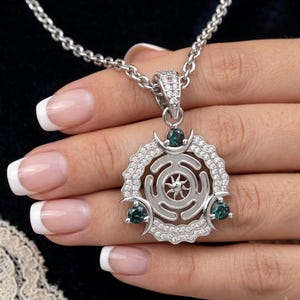 May include: Silver pendant necklace featuring a circular design with a sunburst center, surrounded by clear crystals. The pendant has three teal-colored gemstones and hangs from a silver chain. The necklace is displayed against a dark background.