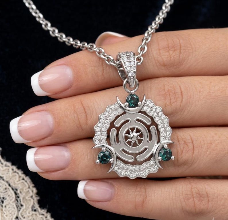 May include: Silver pendant necklace featuring a circular design with a sunburst center, surrounded by clear crystals. The pendant has three teal-colored gemstones and hangs from a silver chain. The necklace is displayed against a dark background.