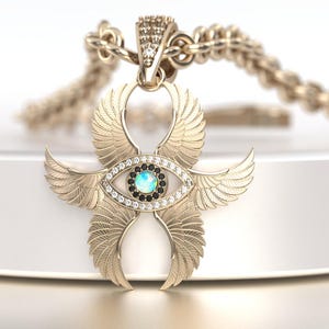 May include: A gold-colored pendant necklace featuring a stylized eye with wings. The eye has a blue center, surrounded by black and clear stones. The wings are detailed with feather patterns. The necklace chain is also gold-colored.