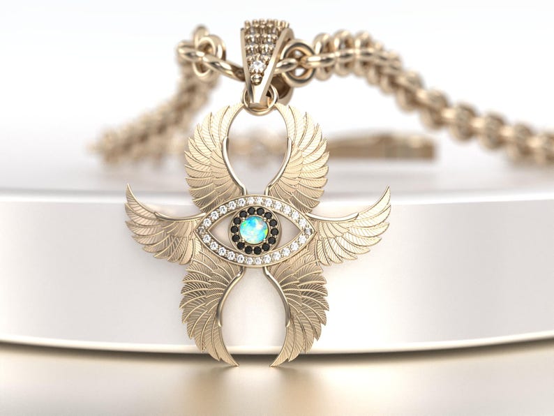 May include: A gold-colored pendant necklace featuring a stylized eye with wings. The eye has a blue center, surrounded by black and clear stones. The wings are detailed with feather patterns. The necklace chain is also gold-colored.