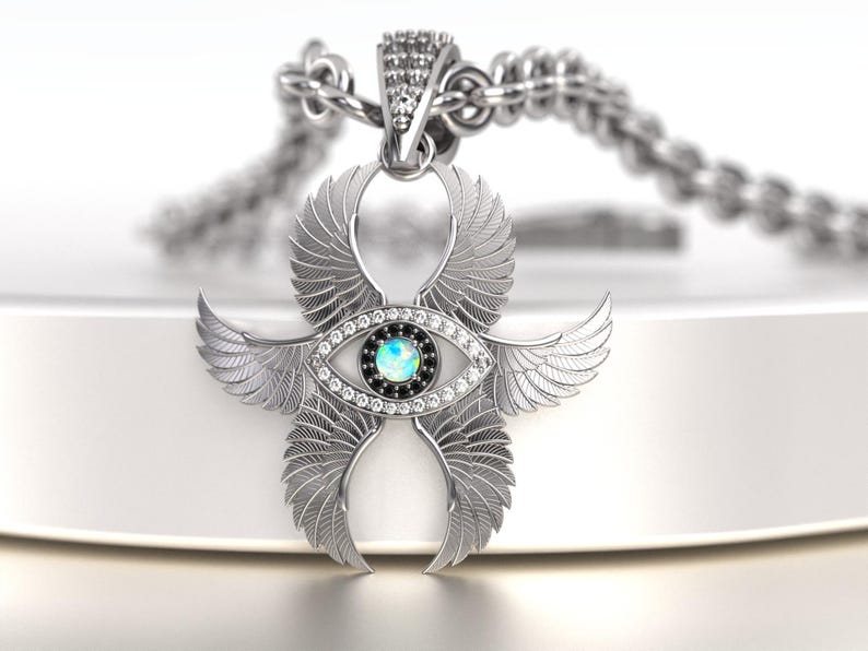 May include: A silver pendant necklace featuring a stylized eye design with feathered wings. The eye has a blue center and is surrounded by clear crystals and a black border. The pendant hangs from a silver chain.