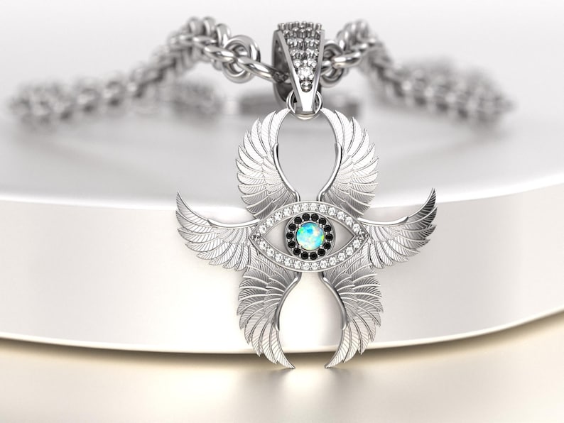 May include: A silver pendant necklace featuring a stylized eye design with wings. The eye has a central opal-like stone surrounded by black and clear stones. The pendant is suspended from a silver chain.