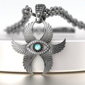 May include: A silver pendant necklace featuring an eye design with wings. The pendant has a central blue-green stone surrounded by clear and black stones. The wings are detailed with feather patterns, and the chain is also silver.