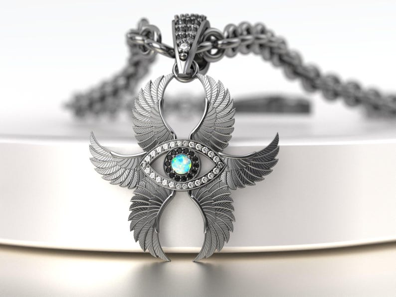 May include: A silver pendant necklace featuring an eye design with wings. The pendant has a central blue-green stone surrounded by clear and black stones. The wings are detailed with feather patterns, and the chain is also silver.