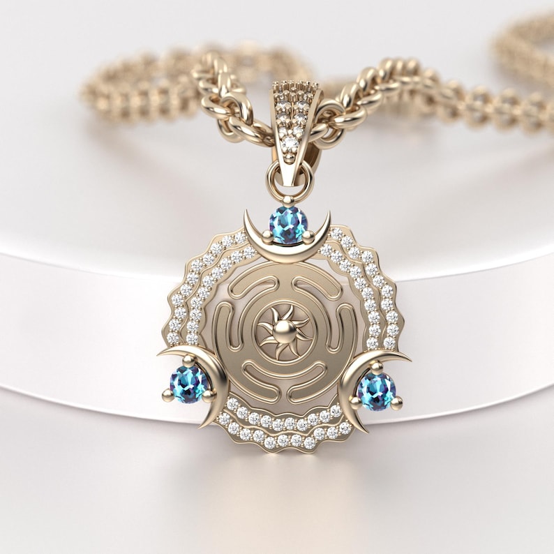 May include: Gold-toned pendant necklace featuring a circular design with a sunburst center and crescent moon accents. The pendant is embellished with small, clear gemstones and three blue gemstones. The necklace chain is also gold-toned.