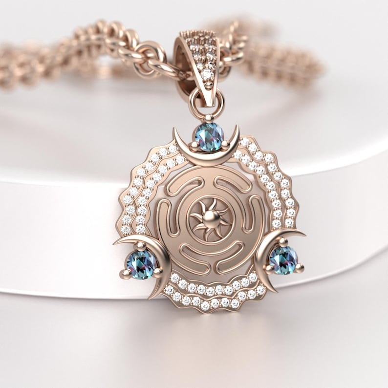 May include: Rose gold pendant necklace featuring a circular design with a sunburst center and three crescent moon accents, each holding a blue gemstone. The pendant is embellished with small, sparkling crystals.