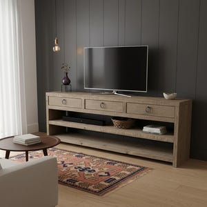 May include: A light-colored wooden TV console with three drawers and open shelving. The console has metal ring pulls and a distressed finish. A large flat-screen TV sits on top. A rug with a geometric pattern is on the floor.
