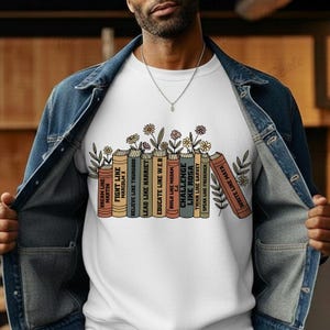 May include: White crew neck sweatshirt with a design of illustrated books and flowers. The book spines display motivational phrases such as "Dream Like Martin" and "Write Like Maya". A casual, comfortable style of sweatshirt.