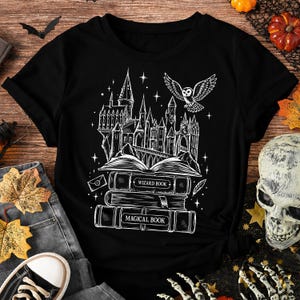 May include: A black t-shirt featuring a white graphic print of a castle perched atop a stack of books. The books are labelled WIZARD BOOK and MAGICAL BOOK. An owl is flying above the castle, surrounded by stars, a small envelope, and a feather.